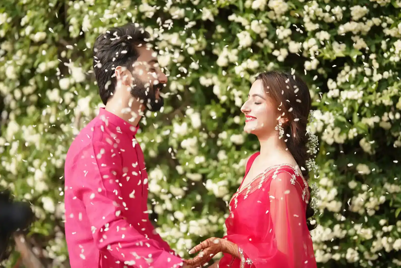 Couple smiling as flower petals fall around them outdoors.